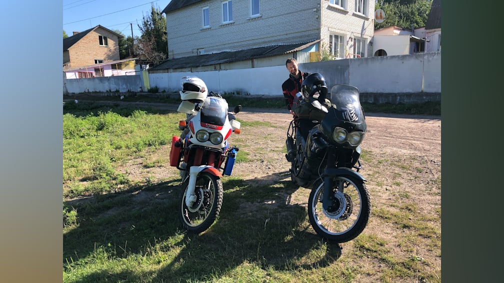 A man is standing next to two motorcycles, one of which is red and white.