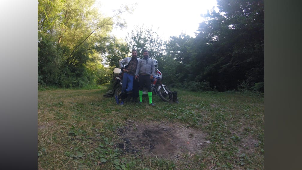Two people on a motorcycle in the woods, one with a green sock on their left leg.