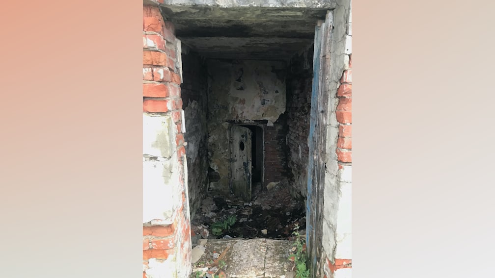 A dilapidated room with a broken door and debris on the ground.