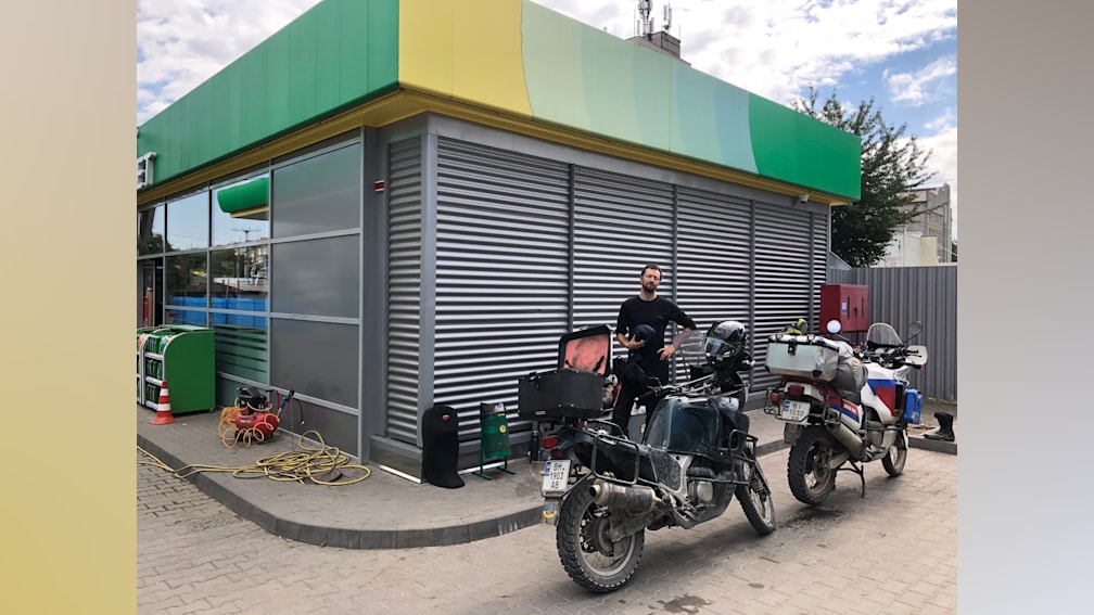 A man stands next to his motorcycle outside a gas station.