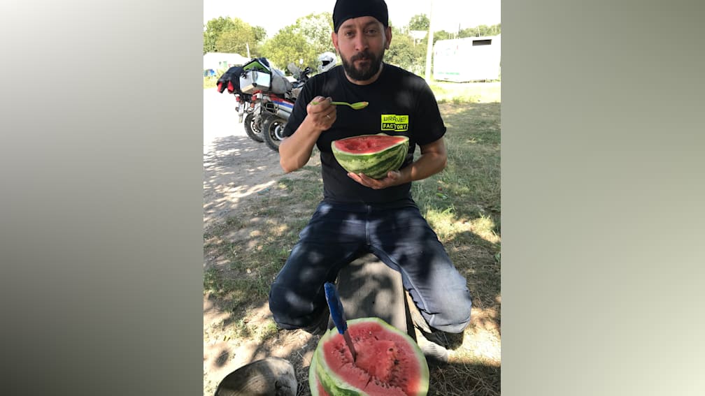A man is sitting outside and eating a watermelon with a yellow spoon in his mouth.