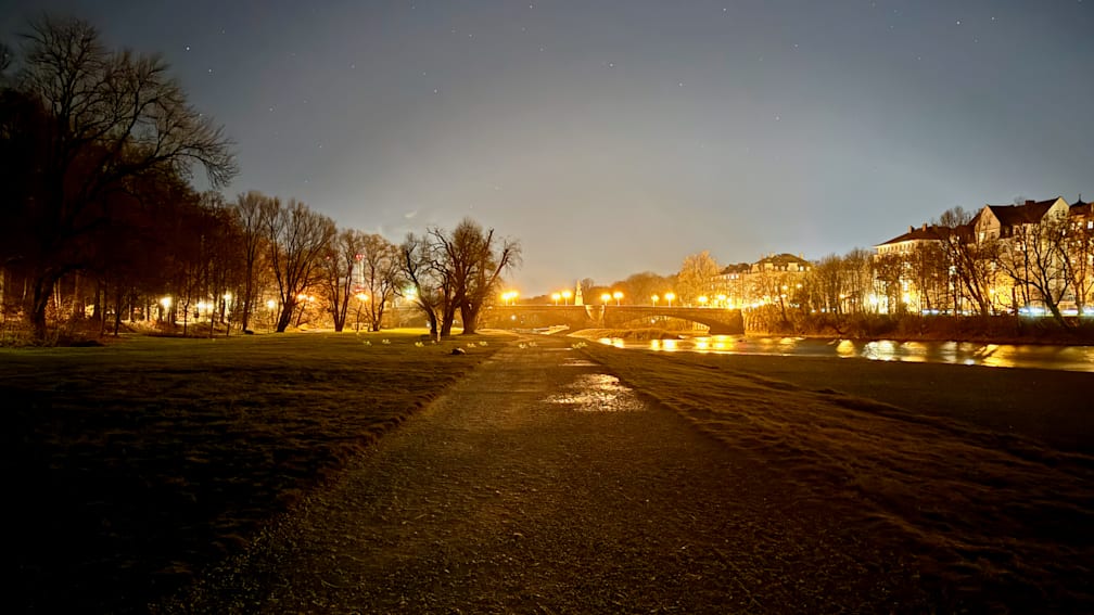 A dark, wet, gravel road is shown with lights from buildings and trees in the background.