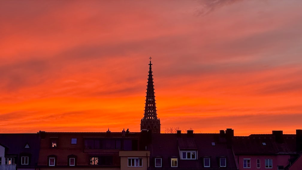 A tall tower with a spire and a cross is silhouetted against a sunset.