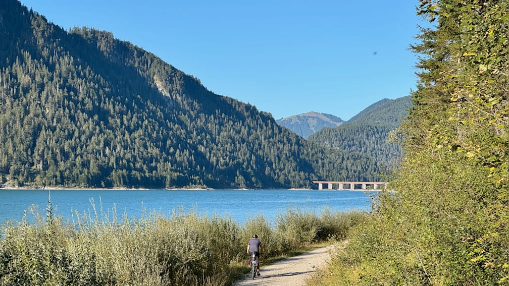 A man on a bike rides along a path near a lake and a bridge.