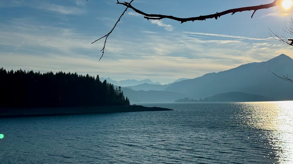 A tree branch with a few leaves is in front of a body of water with a very cloudy sky.