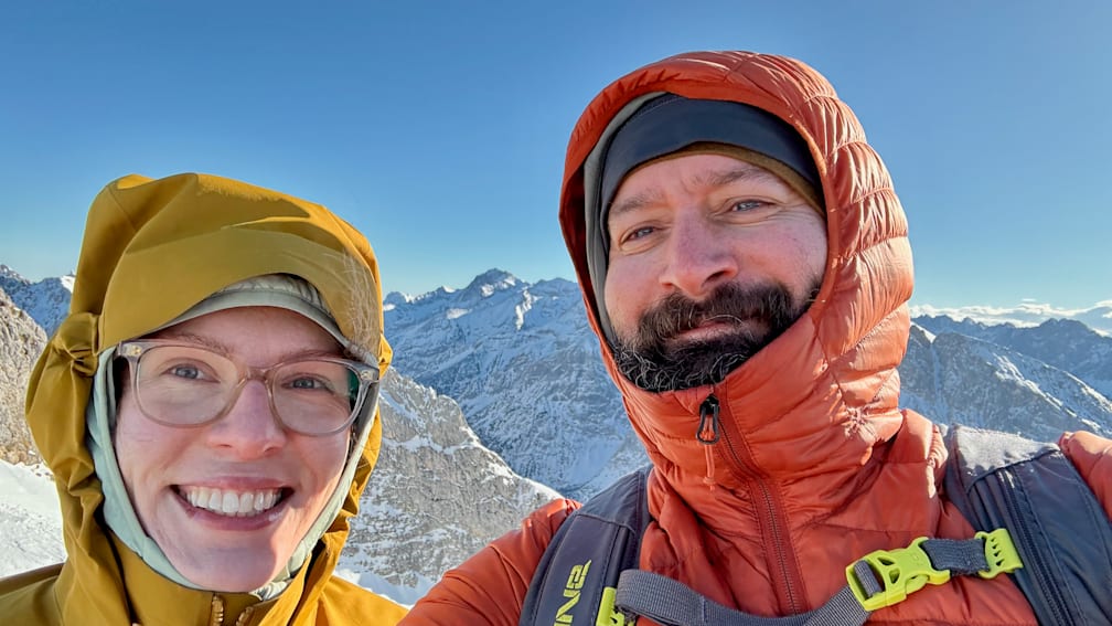 A man and a woman wearing warm coats and taking a photo together at the top of a mountain.