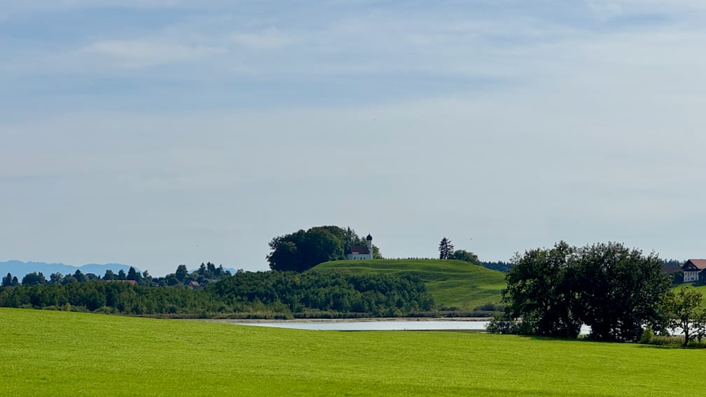 A small white building sits on a green hillside next to a lake.