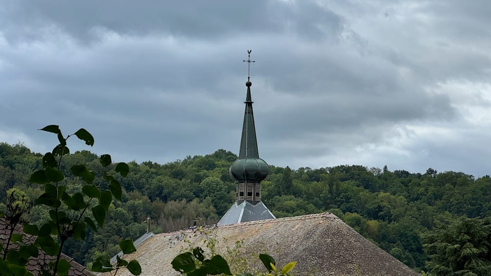 A green steeple with two bells can be seen on a church with a wooded hill in the background.