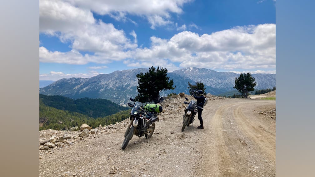Two motorbike riders on a dirt road with mountains in the background.