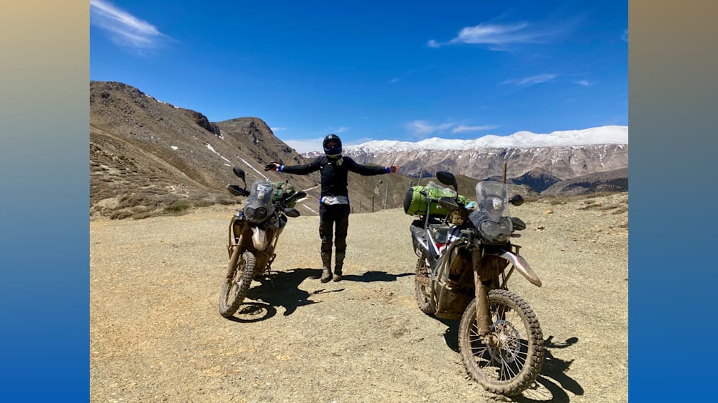 A man standing between two motorcycles in the dirt with his arms up.
