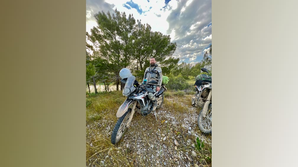 A man on a motorcycle in the woods with a beard and green backpack.