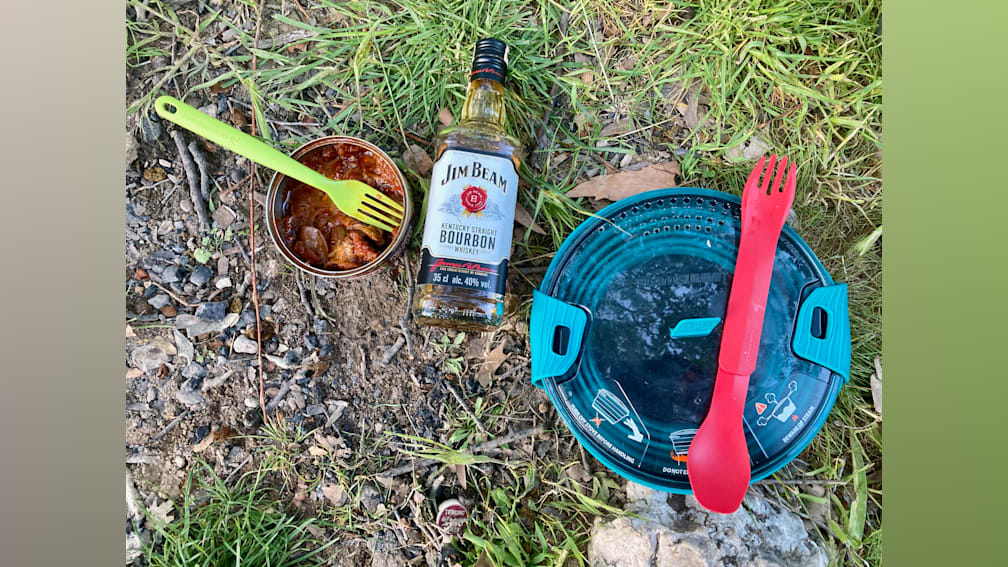 A bottle of Jim Beam is on a rocky area with a fork and spoon on the ground next to it.
