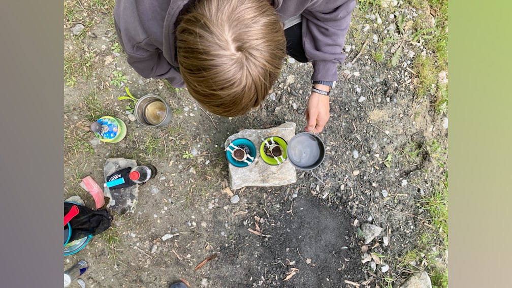 A person is making coffee over a campfire using a camping pot.
