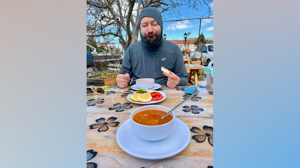 A man with a beard and a grey shirt is sitting at a table eating soup and bread.