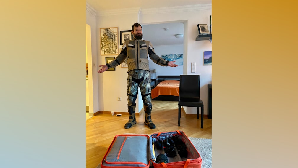 A man in camouflage gear stands in front of a bed with his arms wide open.