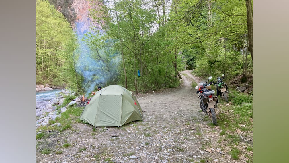 A tent is set up in a wooded area with a motorcycle parked nearby.