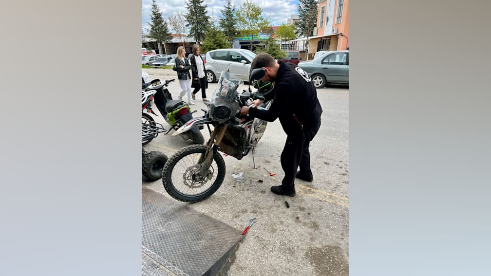 A man works on a motorcycle in a parking lot, with other vehicles around him.