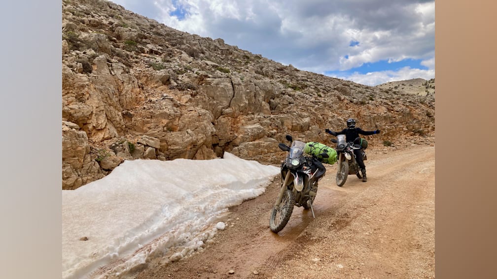 A man is riding a motorcycle through snow on a road with another man on a motorcycle behind him.
