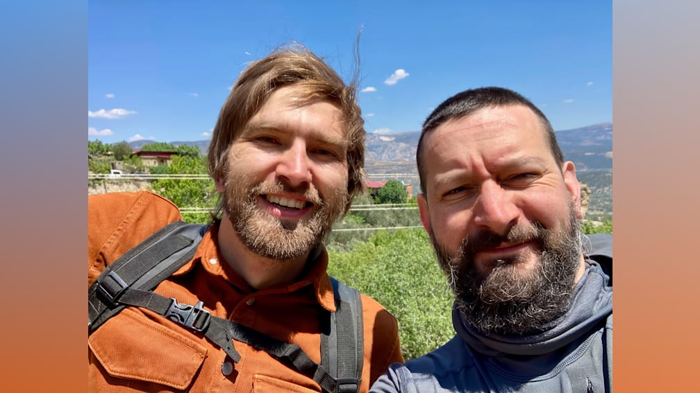 Two men with backpacks, one is smiling at the camera and the other is frowning.