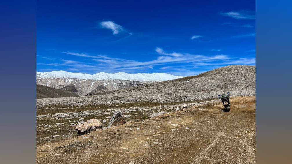 A dirt road winds through a rocky, mountainous pass with a green truck in the foreground.