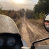 A dirt road has a gauge on a motorcycle in front of a mirror that is being used by another motorcycle rider.
