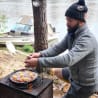 A man cooking outdoors with an even thought of food in a box on a grill.
