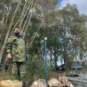 A man in camouflage clothing stands next to two dogs that are sleeping next to a boat.