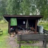 A rustic wood shelter with a green cover on a grassy area with a bench nearby.