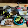 A camping table with a variety of food and utensils, including a blue container with a lid and a pan with food in it.