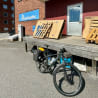 A bicycle is parked on a porch in front of a wood structure and next to a sign that says "pesistertall".