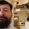 A bearded man looks at his camera as he stands in an airport with a sign that says Assistants.