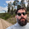 A man wearing sunglasses and a grey shirt, with dirt on his clothes, taking a selfie.