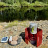 A camping stove sits on some rocks next to some food.