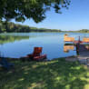A red boat is in the water next to a dock and a chair on the shore.