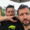 Two men are holding cell phones and one of them is wearing a black t-shirt with a logo that says "grave factory".