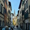 A narrow, brick city street with several cars parked on both sides and a tall, spire-like building in the background.