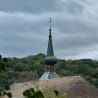 A green steeple with two bells can be seen on a church with a wooded hill in the background.