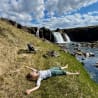 A woman is laying on the grass next to a body of water, with her back to the camera.
