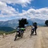 Two motorbike riders on a dirt road with mountains in the background.