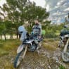 A man on a motorcycle in the woods with a beard and green backpack.