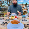 A man with a beard and a grey shirt is sitting at a table eating soup and bread.