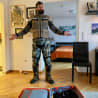 A man in camouflage gear stands in front of a bed with his arms wide open.