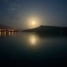 A full moon is reflected on the water and mountains in the distance.