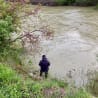 A man in a blue hoodie stands at the edge of a river with his fishing pole.