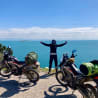 A man on a motorcycle with two other motorcycles next to him, with the ocean in the background.