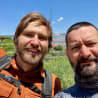 Two men with backpacks, one is smiling at the camera and the other is frowning.