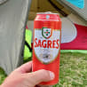 A hand holding a can of Sagres beer in front of a tent.