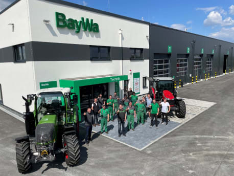 Gruppe von 16 Männern und Frauen vor dem neuen Werkstatt-Gebäude neben einem Fendt-Traktor winken in die Kamera