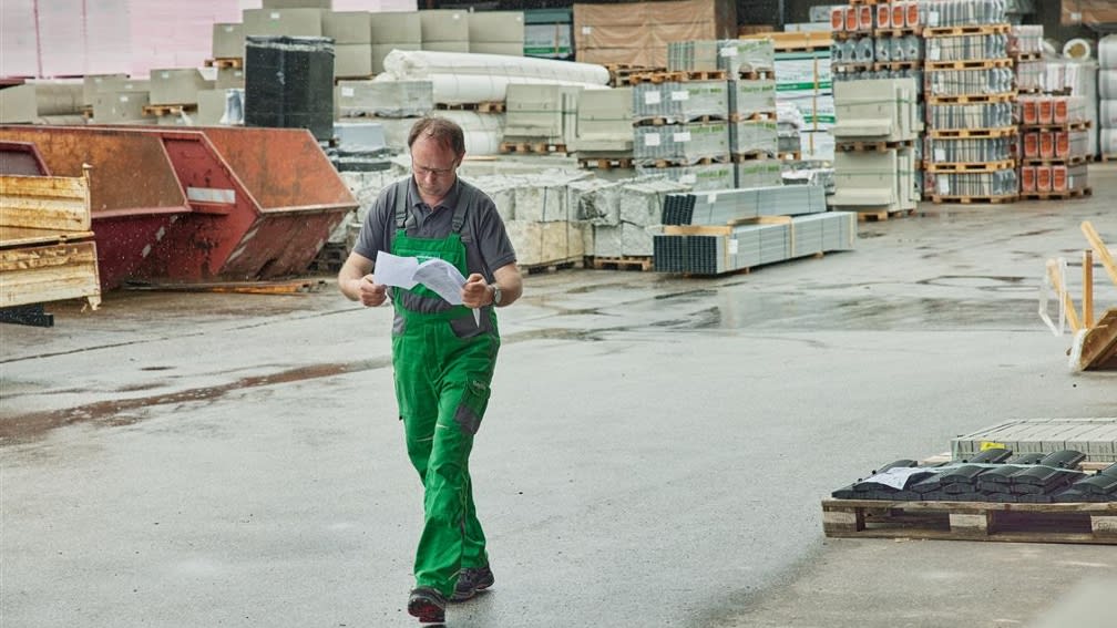 Lagermeister Christian Stinglwagner geht über den Hof seines Lagers bei der BayWa Bau. Er trägt grüne Arbeitskleidung und blättert in Dokumenten.