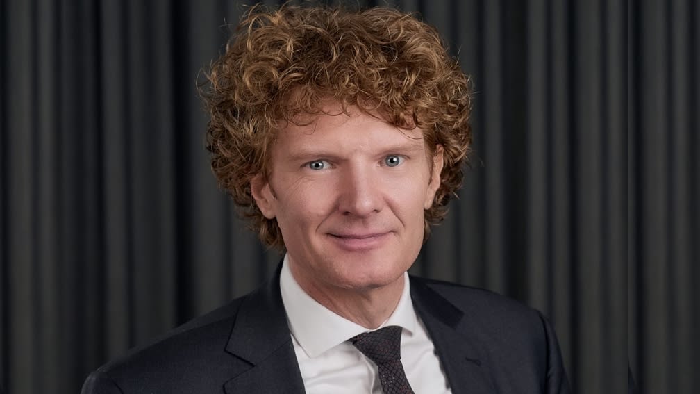 Porträtbild von einem Mann mit lockigem, rotem Haar in weißem Hemd, dunklen Anzug und mit Krawatte vor schwarzem Hintergrund